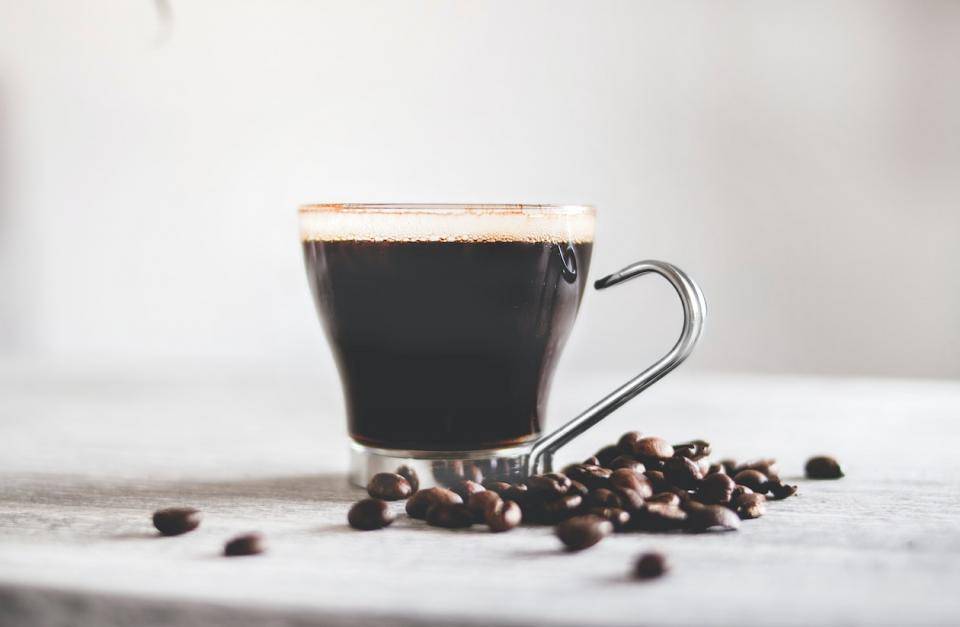 Coffee in a glass cup with handle on white tabletop with white background, coffee beans spilled at base of cup