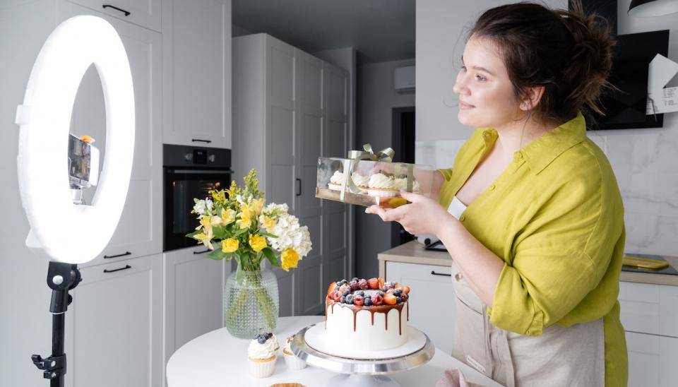 A smiling woman in an apron poses with confections in front of a ring light