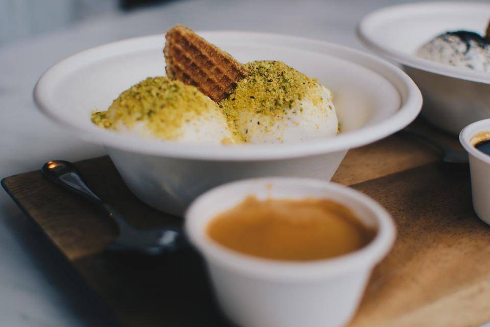 Two scoops of ice cream dusted with pistachio powder with a to-go cup of chocolate sauce