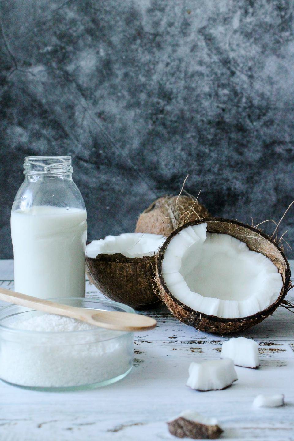 A split coconut, coconut milk in a glass bottle, and a bowl with a wooden spoon