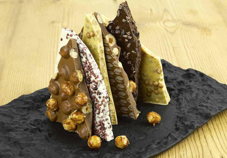 Pieces of chocolate bark standing on end. They are studded with different nuts and displayed on a very textural square black plate