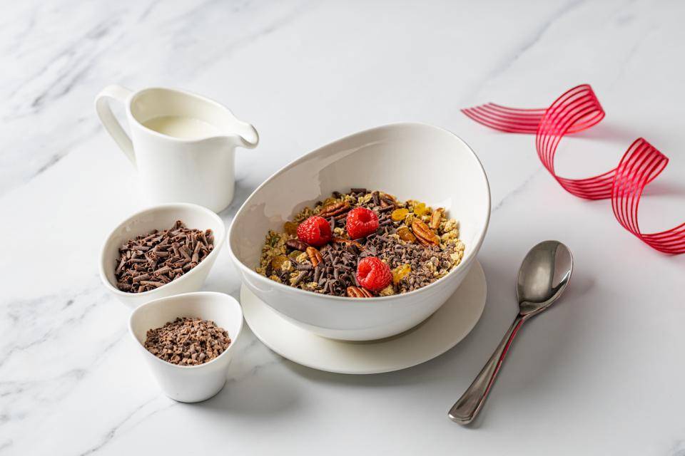 A marble tabletop with a bowl of cereal topped with chocolate shavings and fresh raspberries. Ceramic containers of chocolate garnishes are next to the bowl
