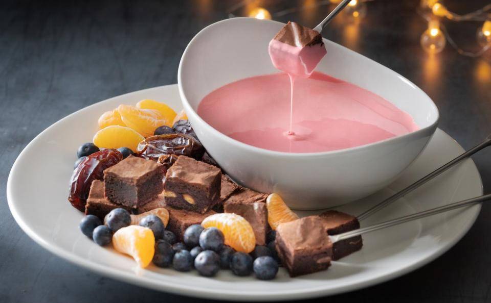 A ceramic bowl of Ruby Chocolate fondue on a plate with brownie chunks and fresh fruit