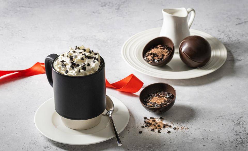 A mug of hot chocolate with whipped cream on a saucer next to a halved hot chocolate bomb with powdered chocolate spilling out