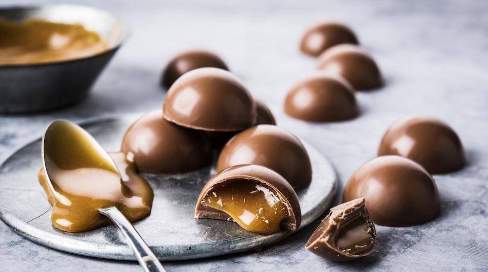  Bombons redondos em um prato e mesa de mármore com uma tigela de calda de caramelo ao fundo, colher de calda de caramelo derramada na colher e saindo de bombom cortado