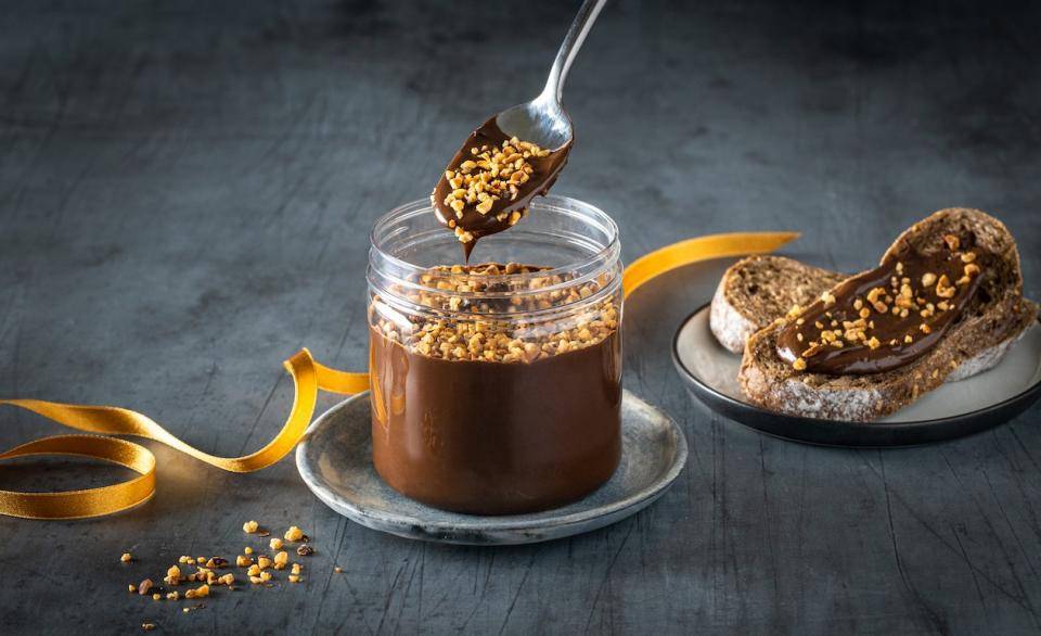 A spoon dips into a jar of chocolate spread topped with nuts