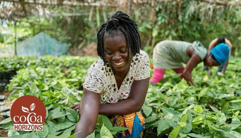 A smiling woman bends down, working in a field of plants, another woman in background, Cocoa Horizons logo in corner