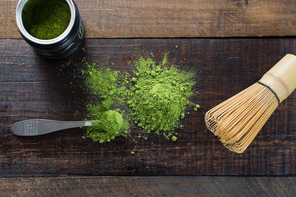 matcha container, whisk on wood background with teaspoon of matcha powder in a small pile of matcha powder