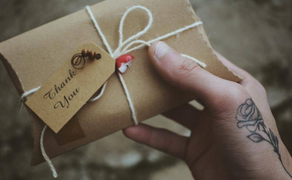 Tattooed hand holds a small package tied with a twine bow, a small tag reading "thank you" is attached