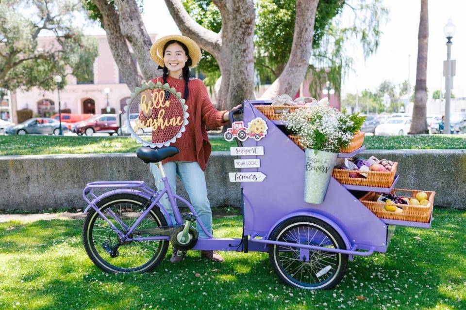 A woman poses in front of a bicycle-style produce/flower cart