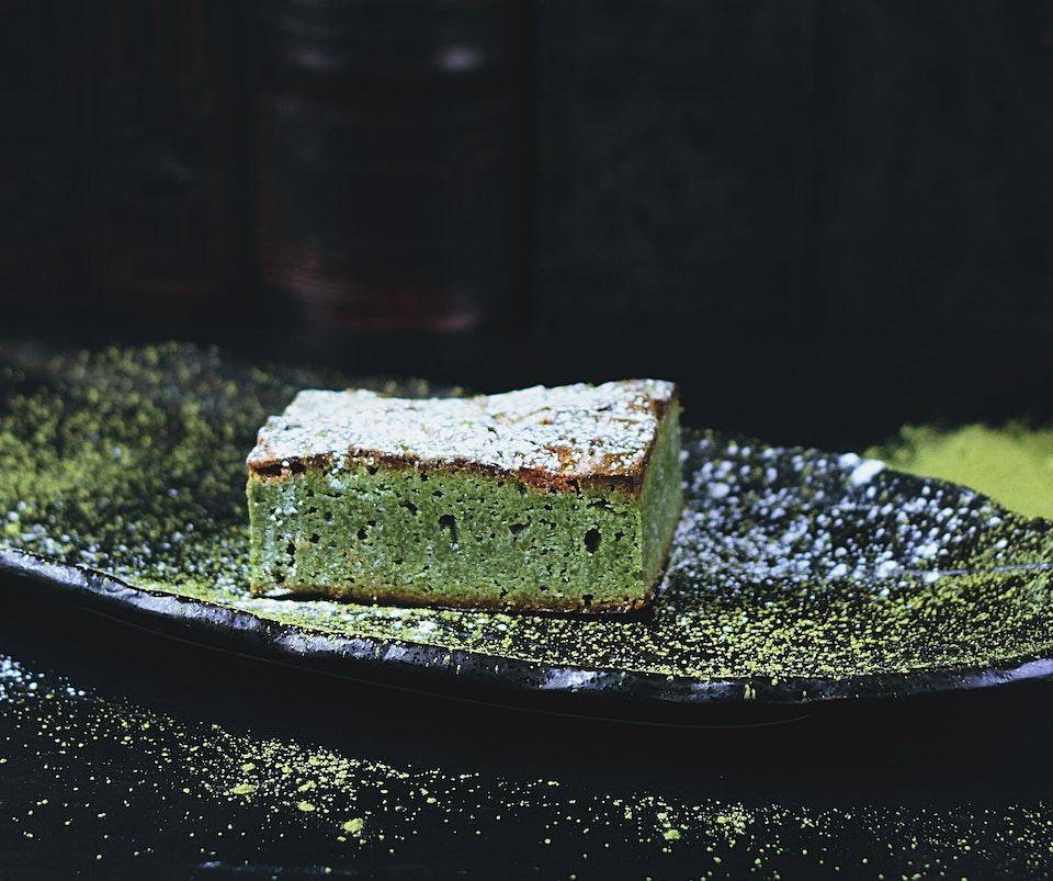 A matcha brownie dusted with powdered sugar on a black stone plate dusted with matcha powder