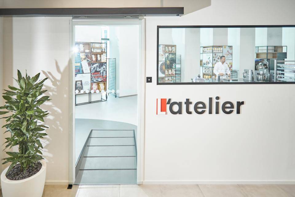 A Chocolate Academy™ Chef can be seen working in the kitchen through the window. Text on wall: l'atelier