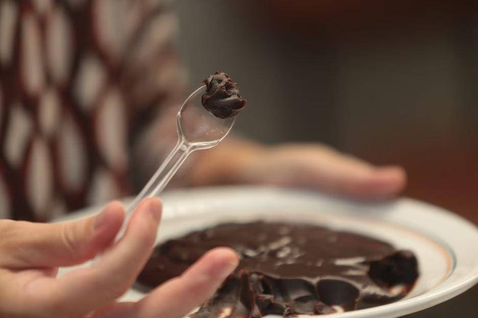 mão segurando em pequena colher de plástico com um pouco de brigadeiro no topo e mão ao fundo segurando prato com brigadeiro