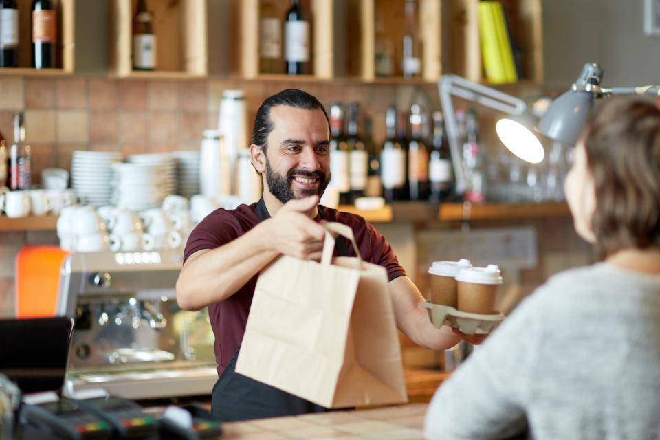 A smiling restaurant employee hands two takeout bags across the counter to a customer