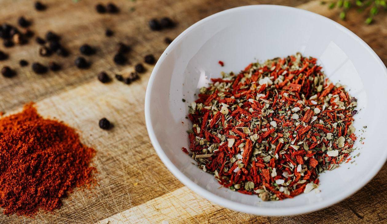 A white bowl of colorful seeds and spices on a wooden table strewn with peppercorns and a pile of dark red spice powder