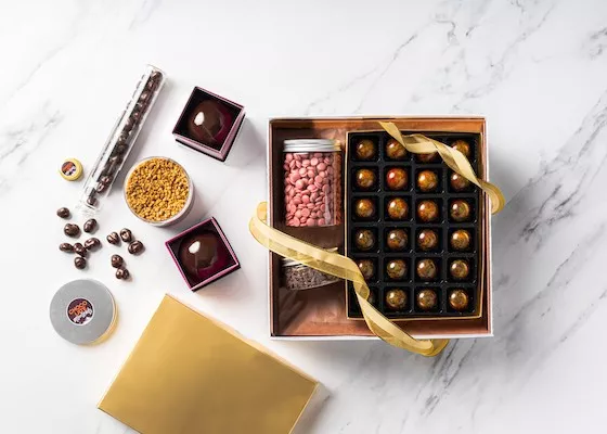 Top-down shot of a marble tabletop with a chocolate box holding bonbons and jars of chocolate garnishes, assorted chocolate items spill attractively around the box