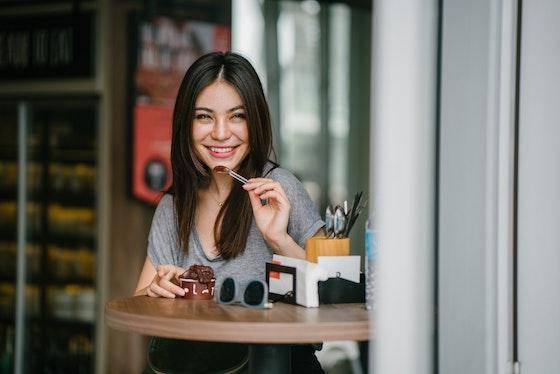 A smiling woman with long, dark hair pauses mid-bite while enjoying a chocolate snack